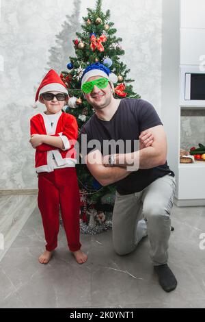 A boy in a Santa suit and his dad in funny sunglasses are on one knee near the Christmas tree with their arms crossed on their chest. Games for Christ Stock Photo