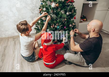 Family - mom, dad and son in a Santa costume are sitting under the Christmas tree on the floor in their apartment and decorating the Christmas tree. Stock Photo