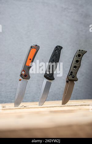 Modern folding knives are stuck with blades in a wooden surface. Various pocket knives. Light background. Stock Photo