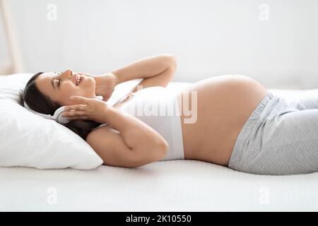 Joyful pregnant woman lying on bed, listening to music Stock Photo