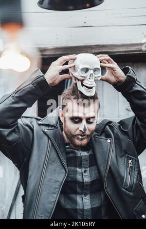 Scary man with dreadlocks looking at skull. Celebrating halloween. Terrifying black white face monster makeup and stylish costume, image.Horror,fun at Stock Photo