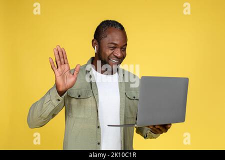 Cheerful adult black male in wireless headphones waving hand, look at laptop, have video call Stock Photo