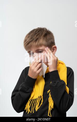 A child blows his nose into a napkin, children's seasonal diseases, a boy wrapped in a scarf, symptoms of the disease. Stock Photo