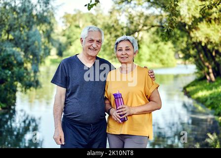 Attractive older couple in sportswear hugging pose look at camera standing in summer park near lake after morning stroll or sport active lifestyle hol Stock Photo