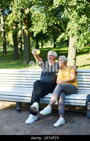 Old spouses make selfie picture or video call sit on bench in summer park. Grey-haired couple look at cell phone screen make videocall outdoor use mod Stock Photo