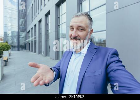 Senior gray-haired handsome man in a suit. Standing near an office building, holding a phone, making a selfie video call, talking, pointing, smiling. Stock Photo