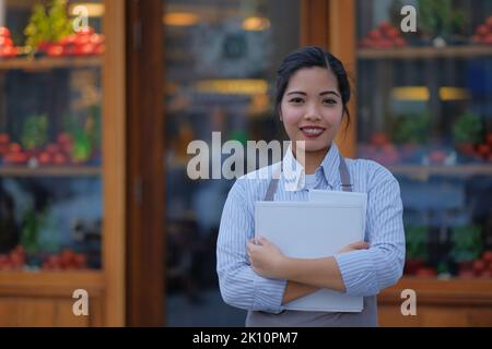 Young Filipino waitress waits outside an Italian restaurant window with menus and a smile. Outdoor portrait of an Asian female in the service industry. Stock Photo