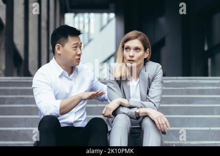 Interracial conflict at work. A young woman and a young asian man are sitting on the steps in front of an office and arguing. A man shouts and proves something to a woman. The woman listens tiredly. Stock Photo