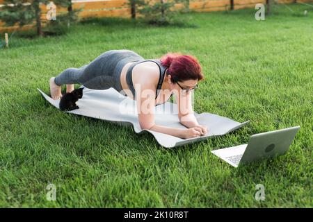 Young plus size woman in sporty top and leggings standing in plank on yoga  mat spending time on green grass in yard. Black kitten walks around her.  Well being and fitness concept 17772580 Stock Photo at Vecteezy