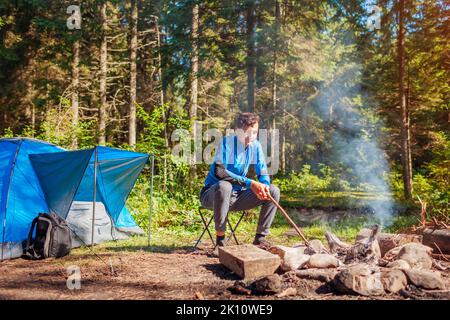 Man traveler relaxing by campfire in forest sitting next to tent checking fire. Summer recreational camping. Traveling alone enjoying nature Stock Photo
