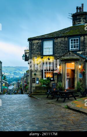 Haworth Main Street (steep hill, old buildings, blue hour evening light, historic Bronte sisters' village, Grade 2 pub) - West Yorkshire, England, UK. Stock Photo