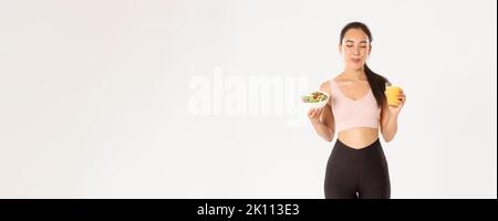 Sport, wellbeing and active lifestyle concept. Smiling cute fitness asian girl in activewear, licking lips satisfied with delicious healthy salad and Stock Photo