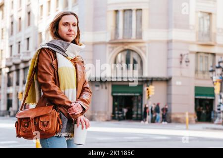 horizontal portrait of a trendy blonde woman waiting public transportation in the street. She holds a reusable coffee mug Stock Photo