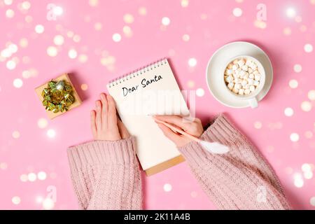 Letter to Dear Santa. Hand holding pink pen decorated with feather and writing in Notepad letter to Santa Claus on pink background with cup of hot Stock Photo