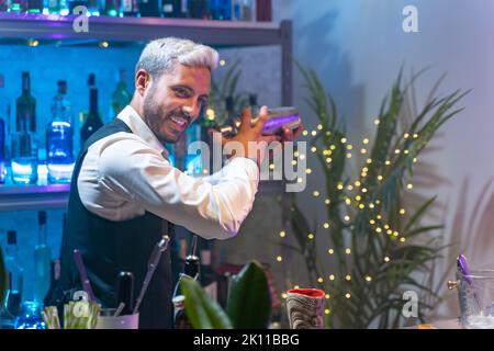Professional bartender in a white shirt and black apron holds a steel shaker in his hands. Blurred bottles on shelves in background Stock Photo