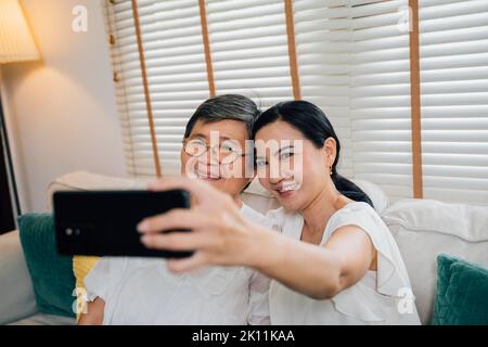 High angle of happy Asian daughter smiling and hugging aged mother while sitting on couch and taking selfie via smartphone together at home Stock Photo
