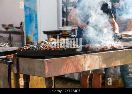 Defocus man cooking bbq meat at festival outdoor. Seafood paella. Chef grilling sausages in park outside. Concept of summer party with families and Stock Photo