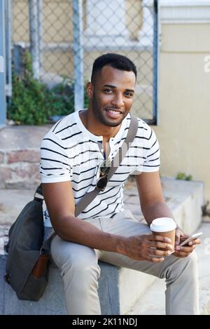 Coffee break on the go. A handsome casually dressed african american businessman sitting in the city and using his mobile while drinking coffee. Stock Photo