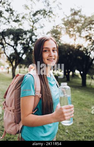Teenage girl wearing turquoise t shirt and a pink backpack, holding a glass bottle with water in nature Stock Photo