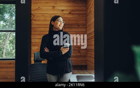 Happy young businesswoman looking away with a smile while holding a smartphone. Cheerful female entrepreneur working remotely in a modern co-working o Stock Photo