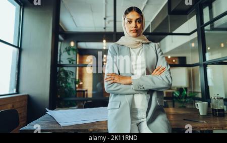 Confident Muslim businesswoman looking at the camera while standing with her arms crossed. Young female entrepreneur wearing a hijab in a modern offic Stock Photo