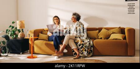 Two businesswomen having a discussion while working on a laptop in a lobby. Successful female entrepreneurs sitting on a couch in a modern coworking o Stock Photo
