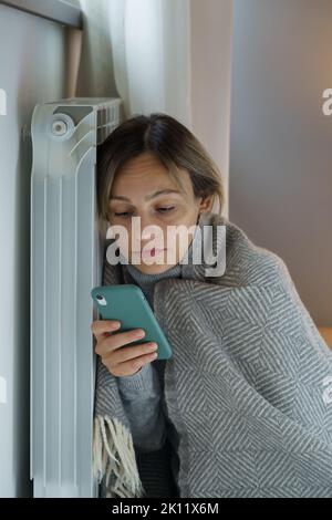 Upset woman leaning on heating radiator searches vacancy for applying for job scrolling smartphone Stock Photo