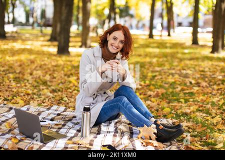 Cheerful caucasian young red-haired female in raincoat enjoys favorite hot drink, uses laptop, sits on plaid in park Stock Photo