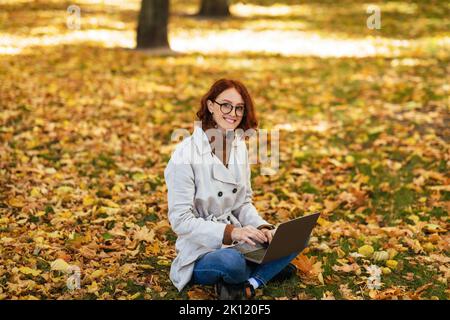 Cheerful caucasian millennial red-haired lady in raincoat and glasses typing on laptop, sit on ground Stock Photo