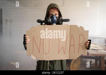 No war concept. Angry woman in protective uniform and gas mask after apocalypse holds a sign No War screaming furiously in a building background. Stock Photo