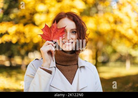Portrait of glad pretty caucasian young red-haired lady in raincoat puts red leaf to eye, enjoys freedom Stock Photo