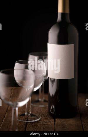 A bottle of red wine with an empty white label and three empty small wine glasses on a wooden table Stock Photo