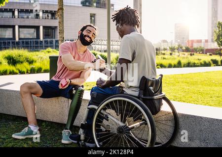 An hispanic man with an artificial prosthesis on his leg shakes hands with his African friend sitting in a wheelchair - diversity lifestyle concept - Stock Photo
