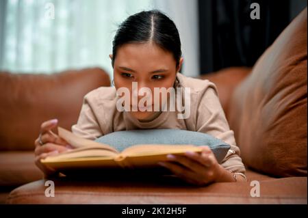 Attractive and gorgeous young Asian female concentrating on reading a book while relaxing, lying on her comfortable sofa. Activity and hobby concept Stock Photo