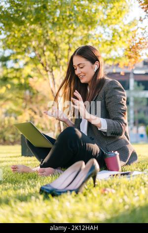 Young caucasian businesswoman having video call conversation online on laptop sitting outdoors in city park on green grass lawn, smiling and gesturing Stock Photo