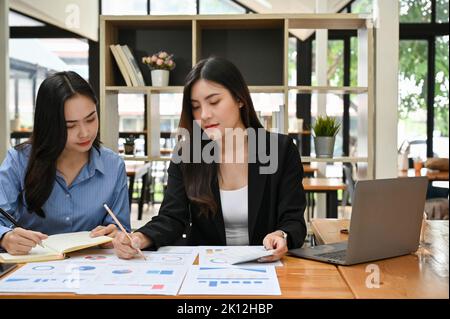 Two attractive young Asian businesswomen working and analyzing business financial data on the report together. Stock Photo