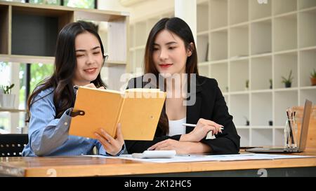 Two beautiful young Asian businesswomen coworkers, working together in the office, looking for information in a book. Stock Photo