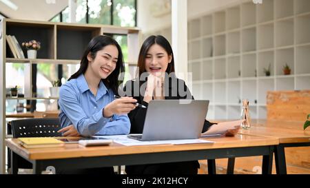 Happy and cheerful young Asian businesswomen or female business colleagues are watching something funny on laptop together during the lunch break. Stock Photo