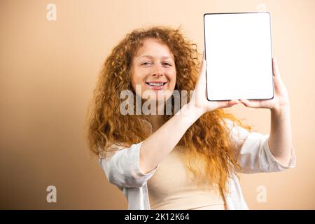 Close up photo of a pretty young redhead woman smiling widely holding a tablet with shiny screen in her hands. Stock Photo