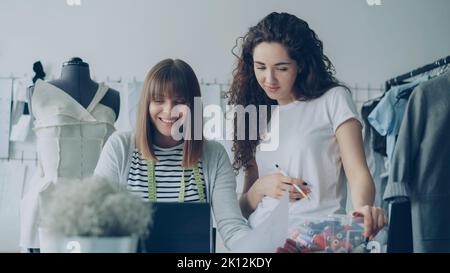 Cheerful creative clothing designers are watching laptop screen, having friendly conversation, gesturing, smiling and laughing together in light workshop. Fun at workplace concept. Stock Photo