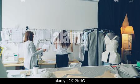 Young female entrepreneurs clothing designers standing in front of wall with hanging sketches, discussing drawings and placing images with clips. Creative team working together concept. Stock Photo
