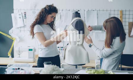 Creative fashion designers are pinning cut out pieces of fabric to mannequin while sewing women's garment in modern light studio. Ladies are concentrated on their work. Stock Photo