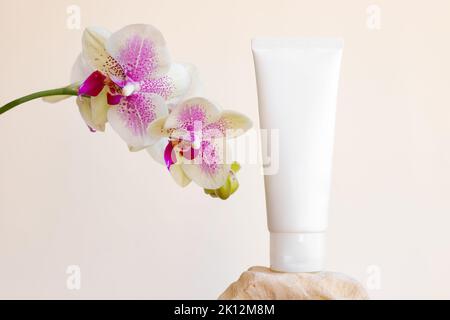 Mockup white squeeze bottle cosmetic tube on rock, phalaenopsis orchid flowers on beige background. Body cream, gel, skin care, sunscreen, moisturizer Stock Photo