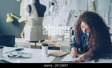 Young female artist fashion designer is drawing women's garment sketch with pencil at table in modern studio. Mannequin, sewing items, take-away coffee are visible. Stock Photo