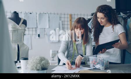 Female employee of tailor shop is measuring garment drawings with sewing threads while her attractive colleague is showing her tablet screen and talking to her. Stock Photo