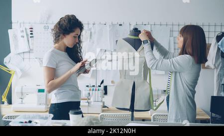 Clothing designer entrepreneurs are measuring mannequin parts while working together in nice modern studio. Young attractive woman is using tablet to enter measurement results. Stock Photo