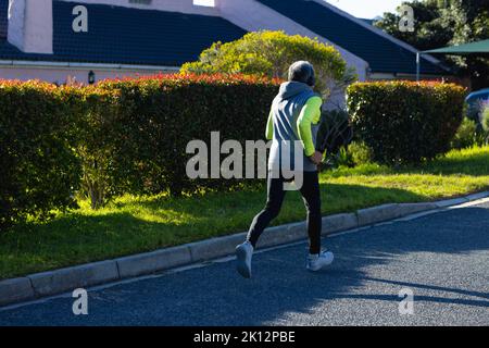 Rear view of senior biracial man in sports clothes and headphones running in road Stock Photo