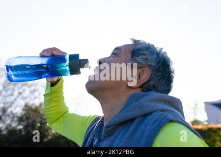 Thirsty senior biracial man in sports clothes drinking water while out jogging Stock Photo