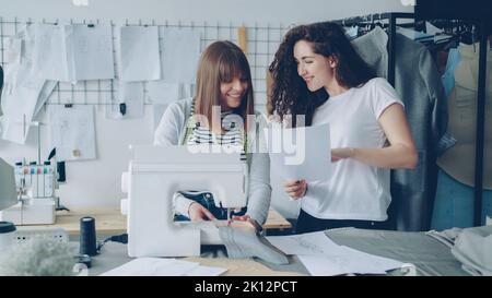 Professional seamstress is stitching fabric with sewing machine then adjustig equipment while her female colleague is showing her sketches. Light studio with tailoring items in background. Stock Photo