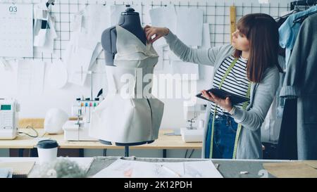 Young female fashion designing entrepreneur is working with tablet while checking clothing pinned to tailoring dummy. Modern technologies in clothes manufacturing concept. Stock Photo